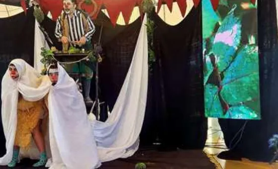 Cena de teatro com atores sob um tecido branco e painel de LED ao lado